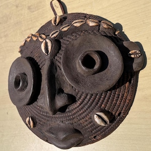 May include: A dark brown, handcrafted mask with a woven texture and large, circular eye and mouth details. The mask is adorned with several small, white seashells and a leather loop for hanging. The mask is likely used for decorative purposes.