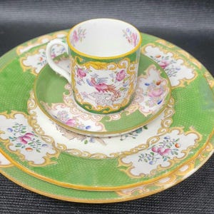 May include: A vintage porcelain tea set comprising a cup, saucer, and two plates. The set features a green background with a textured pattern, gold trim, and floral and bird designs. The cup and plates are stacked.