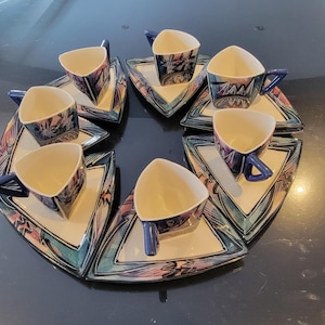 May include: A set of eight ceramic teacups and saucers. The cups are triangular with blue handles and the saucers are triangular with a cream-colored center and a colorful geometric design in shades of blue, pink, and purple.