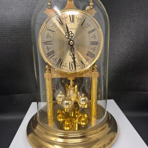 May include: A gold-tone anniversary clock encased in a clear glass dome. The clock features a circular face with black Roman numerals, ornate black hands, and the text "IRKS" and "Quartz" printed on the dial. It has a rotating pendulum base.