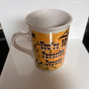 K&ouml;nnte beinhalten: Wei&szlig;er Keramikbecher mit gebogenem Henkel. Der Becher hat ein Design mit Cowboyhut, Sternen und dem Text "You're My Favorite Deputy" in Schwarz. Der Hintergrund ist eine Mischung aus Orange und Braun.