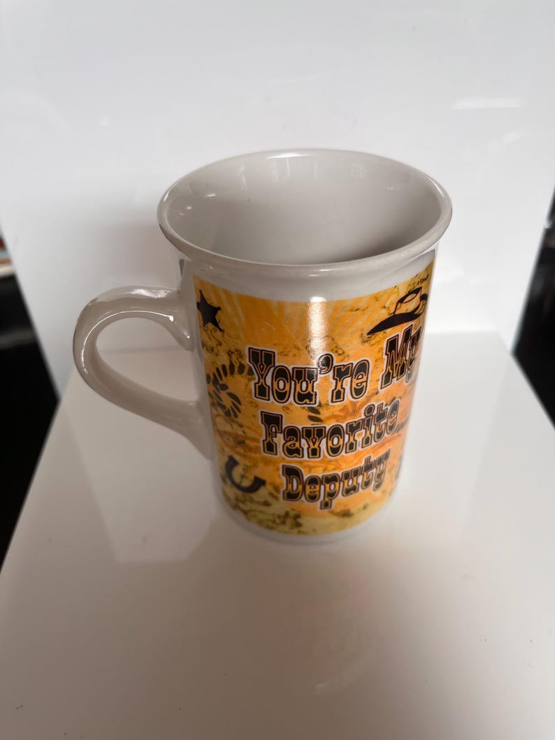 K&ouml;nnte beinhalten: Wei&szlig;er Keramikbecher mit gebogenem Henkel. Der Becher hat ein Design mit Cowboyhut, Sternen und dem Text "You're My Favorite Deputy" in Schwarz. Der Hintergrund ist eine Mischung aus Orange und Braun.