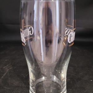 May include: A clear pint glass with the Guinness logo in white and gold. The glass has a curved shape, wider at the top and bottom, and tapers in the middle. The glass is empty.