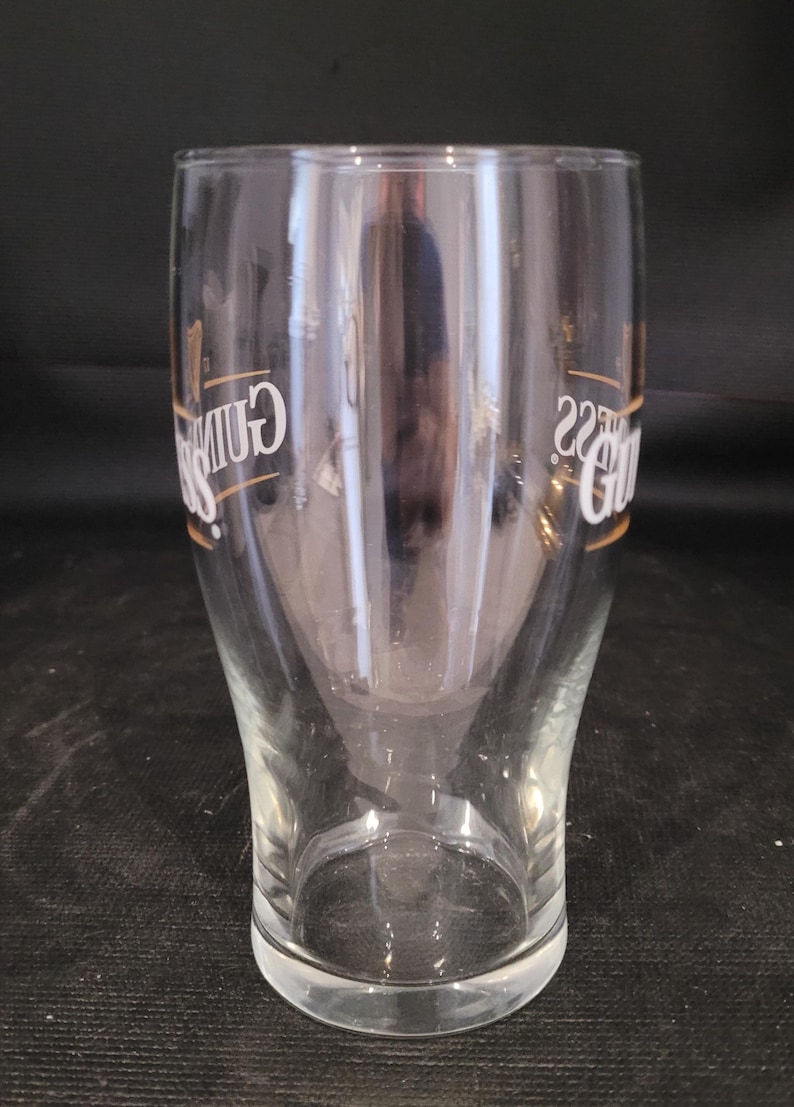 May include: A clear pint glass with the Guinness logo in white and gold. The glass has a curved shape, wider at the top and bottom, and tapers in the middle. The glass is empty.