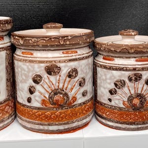 May include: A set of four cylindrical ceramic canisters with lids, featuring a cream-coloured base with brown and orange hand-painted floral patterns and a textured lattice design around the bottom edge, arranged in descending order of size.