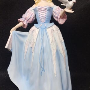 May include: A porcelain figurine of a woman in a blue and pink dress holding a white dove. She is wearing a black hat and has blonde hair.