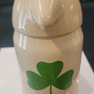 May include: A beige ceramic lidded jar featuring a glossy finish and a prominent green shamrock graphic on the front. The lid is shaped like a stylized bird head with a pointed beak, black circular eyes, and red accents above the eyes.