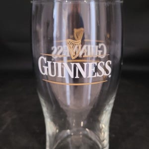 May include: Clear pint glass with the Guinness logo in gold and white. The glass is 6 inches high and 3.25 inches in diameter. The Guinness logo includes a harp symbol and the text "GUINNESS".