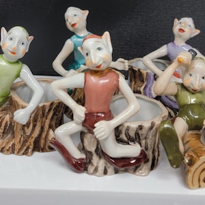 May include: A grouping of five ceramic elf figurines. Each elf is posed in a different position, with varying outfits of green, red, and blue. They are perched on or near tree stump planters, with detailed bark textures.