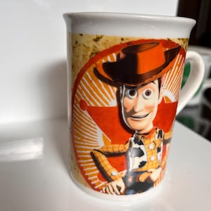K&ouml;nnte beinhalten: Wei&szlig;er Keramikbecher mit einem Cartoon-Bild von Woody aus Toy Story. Der Becher hat einen wei&szlig;en Henkel und ein Design in Orange, Gelb und Braun. Der Becher hat eine Standardgr&ouml;&szlig;e f&uuml;r Hei&szlig;getr&auml;nke.