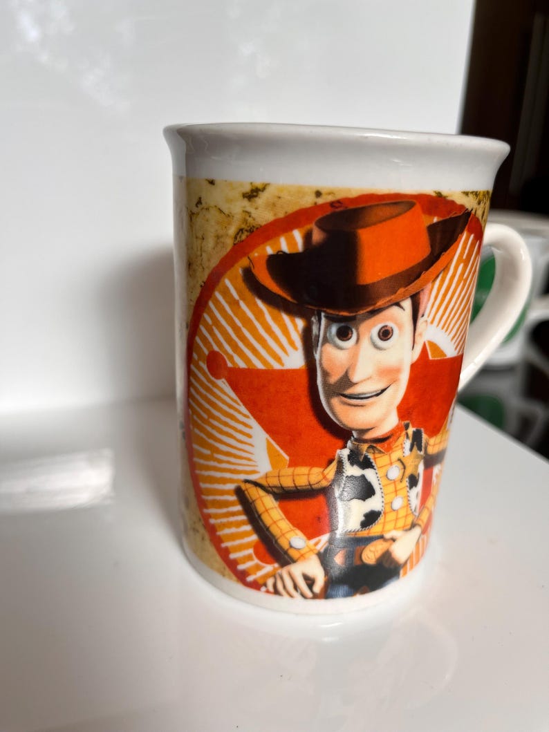 K&ouml;nnte beinhalten: Wei&szlig;er Keramikbecher mit einem Cartoon-Bild von Woody aus Toy Story. Der Becher hat einen wei&szlig;en Henkel und ein Design in Orange, Gelb und Braun. Der Becher hat eine Standardgr&ouml;&szlig;e f&uuml;r Hei&szlig;getr&auml;nke.