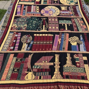 May include: A woven throw blanket with a bookshelf design, displaying books, clocks, and decorative items. The colour scheme includes black, burgundy, gold, and green. The blanket has a fringed edge, suitable for home decor.