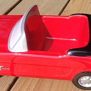 May include: A red ceramic toy car, a convertible, with black and white detailing. The car features a white windscreen frame and black trim. The wheels are black with silver hubcaps. The Mustang logo is visible.