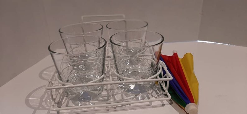 May include: Four clear glass tumblers are stacked in a white wire rack. A small rainbow-coloured umbrella is on the right side of the image.