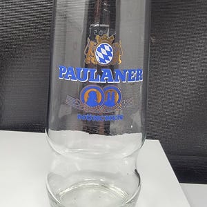 May include: Clear glass beer glass featuring the "PAULANER" logo in blue and gold. The glass has a distinctive shape, wider at the top and bottom, with a narrower central section. The word "MÜNCHEN" is also visible.