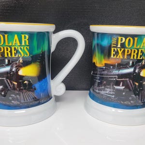 Op de afbeelding: Twee keramische mokken met de tekst "THE POLAR EXPRESS" en een treinillustratie. De mokken hebben een witte basis, een gebogen handvat en een gele rand. De achtergrond heeft een kleurrijk aurora borealis-ontwerp.