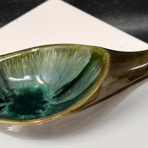 May include: A ceramic dish shaped like a duck, featuring a glossy finish with a deep olive green exterior and a vibrant, variegated teal and dark green interior glaze. The elongated form includes a defined head, neck, and a wide, shallow basin.