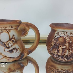 May include: Two ceramic mugs with Australian themes. One mug features a koala holding a joey with the word "Australia" on the top. The other mug has a brown glaze and features a kangaroo and other Australian animals with the words "Aussie Bunch" on the top.
