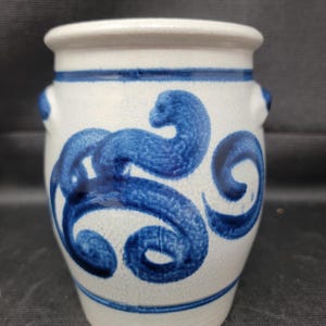 Op de afbeelding: Kleine, gebroken witte keramische vaas met een blauw wervelontwerp. De vaas heeft een afgeronde body, een licht uitlopende rand en twee kleine handvatten. Het blauwe ontwerp is in een vloeiende stijl geschilderd.