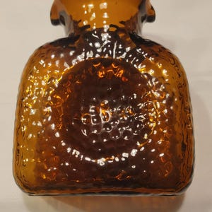 Puede incluir: Botella de vidrio color ámbar con superficie texturizada. La botella tiene un cuerpo redondeado y un cuello acampanado. El vidrio tiene un aspecto martillado o con hoyuelos. La botella es probablemente para uso decorativo.