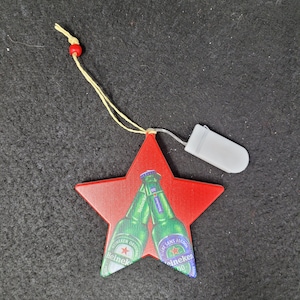 May include: A red star-shaped ornament with two Heineken beer bottles clinking. The ornament has a yellow string with a red bead and a small white battery pack attached.