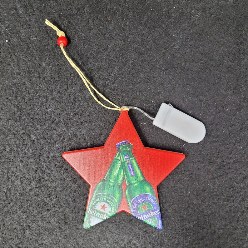 May include: A red star-shaped ornament with two Heineken beer bottles clinking. The ornament has a yellow string with a red bead and a small white battery pack attached.