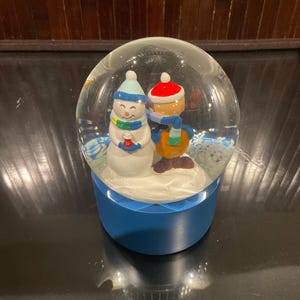 Puede incluir: Un globo de nieve con base azul presenta un muñeco de nieve y un niño con ropa de invierno. El muñeco de nieve lleva un gorro y bufanda azules, mientras que el niño tiene un gorro y bufanda rojos. El globo está lleno de agua y purpurina.