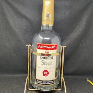 May include: A clear glass bottle of Gooderham's Bonded Stock Canadian Whisky in a gold metal stand. The bottle label features the text "Gooderham's Bonded Stock", "Canadian Whisky Canadian", "Gooderham's", "1832", "Herts", "1.331/3 FL OZ (110 3/9 ML)", "Canada's First Distillery", and "Première Distillerie au Canada".