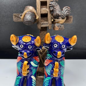 Könnte beinhalten: Zwei blaue Stierfiguren aus Keramik mit goldenen Hörnern und bemalten Blattmustern in Rot, Grün und Weiß. Dahinter steht eine kreuzförmige Holzstruktur, die einen kleinen dunklen Keramiktopf und eine dunkle Hahnfigur hält.
