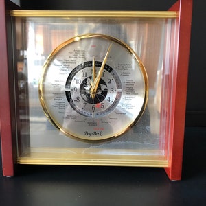 May include: A gold-toned world clock with a black face and a red GMT line. The clock is encased in a clear glass and a wooden frame with a gold trim. The clock face shows the time in various cities around the world. The clock is made by Bey-Berk.