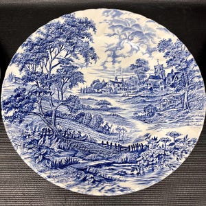 May include: Blue and white ceramic plate depicting a rural landscape. The plate showcases a detailed scene with trees, buildings, and a river, all in a traditional blue and white colour palette. The design is reminiscent of classic English china.