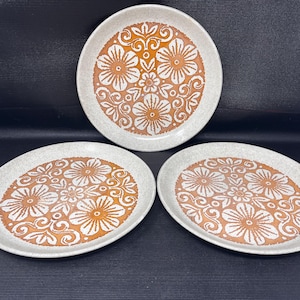 May include: Three round ceramic plates featuring a speckled off-white base with a central orange and white floral pattern. Each plate displays a repeating design of stylized flowers and swirling leaf motifs centered on the surface of the dish.