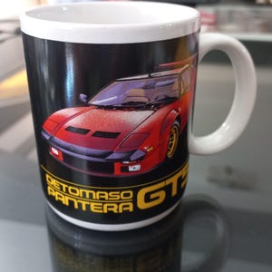 May include: A white ceramic mug with a black and yellow De Tomaso Pantera GT graphic. The car is red and has a black interior. The mug has a white handle.