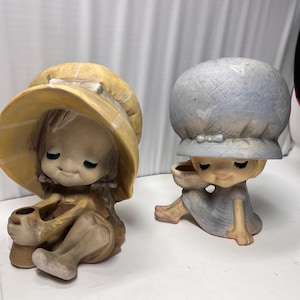 Peut inclure: Deux figurines en céramique d'enfants assis. L'une porte un chapeau jaune et tient un petit pot. L'autre porte un chapeau bleu et tient un panier. Les deux ont les yeux fermés et sont peints dans des tons sourds.
