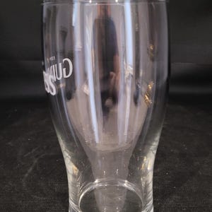 May include: A clear, tulip-shaped pint glass with the Guinness logo in white. The glass has a wide top, a narrow waist, and a sturdy base. The glass is empty and reflects the surrounding environment.