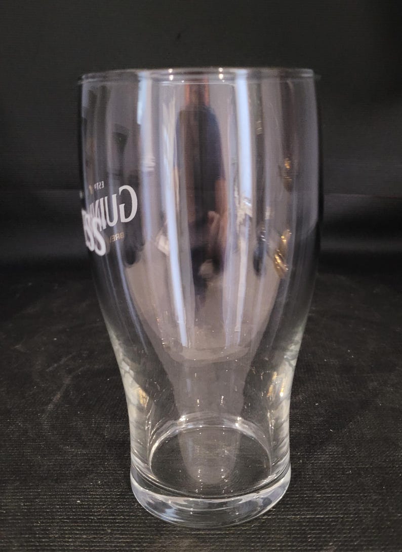 May include: A clear, tulip-shaped pint glass with the Guinness logo in white. The glass has a wide top, a narrow waist, and a sturdy base. The glass is empty and reflects the surrounding environment.