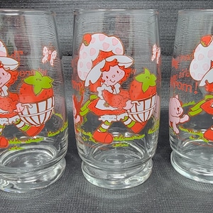 Puede incluir: Tres vasos de vidrio transparente con un diseño de personaje de fresa de dibujos animados. El personaje lleva un sombrero blanco con lunares rojos y un vestido rojo. Los vasos tienen una base ancha y una parte superior ligeramente acampanada.