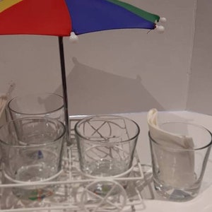 May include: A white metal drink holder with four clear glass cups and a small colourful umbrella. The umbrella has yellow, red, blue and green panels.