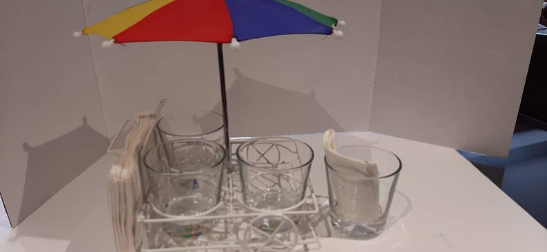 May include: A white metal drink holder with four clear glass cups and a small colourful umbrella. The umbrella has yellow, red, blue and green panels.