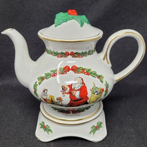 May include: White porcelain teapot with a Christmas scene featuring Santa Claus, a child, and toys. The teapot has a green holly leaf lid with a red berry. The teapot is on a white pedestal base with a gold trim.