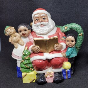 May include: A ceramic teapot shaped like Santa Claus reading a book to two children. Santa is wearing a red suit with white trim and a white beard. The children are wearing colourful clothing. The teapot has a green handle shaped like a Christmas wreath.