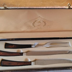 May include: A vintage carving set in a wooden case. The set includes a carving fork and two knives with silver blades and dark wood handles. The case has a cream-colored interior and a decorative emblem.