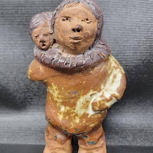 May include: A ceramic sculpture of a person carrying a child on their back. The sculpture is primarily brown with touches of white and a dark brown collar. The figures have simple facial features and are wearing hooded garments. The piece has a rustic, handmade appearance.