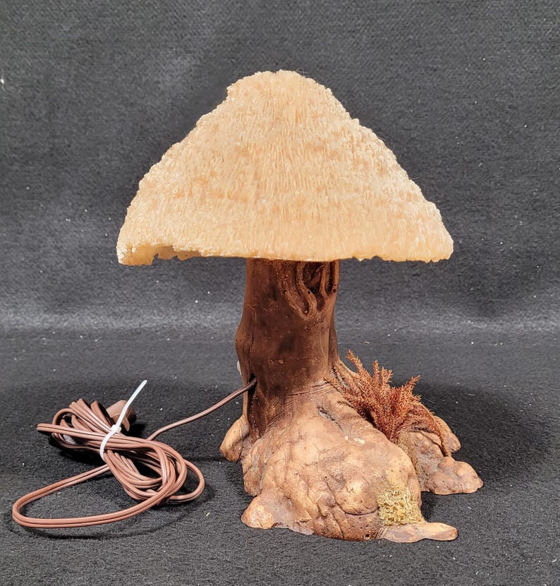 May include: A whimsical mushroom-shaped lamp with a textured, beige lampshade and a brown, tree-trunk-like base. The lamp has a brown power cord. The lamp is designed to resemble a mushroom.
