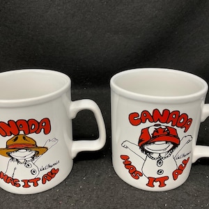 May include: Two white ceramic mugs with red and black illustrations of cartoon figures wearing hats. The mugs have the text "Canada Has It All" on them.