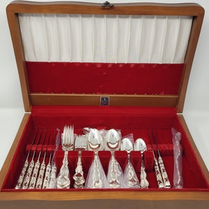 Puede incluir: Una caja de madera con un interior de terciopelo rojo, que contiene un juego de cubiertos de plata. Los cubiertos incluyen cuchillos, tenedores y cucharas, todos con diseños ornamentados.