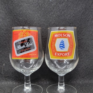 May include: Two clear stemmed beer glasses. One glass has a red and black label that reads "Carling Black Label Bier Beer". The other glass has a yellow and white label that reads "Molson Export Ale".