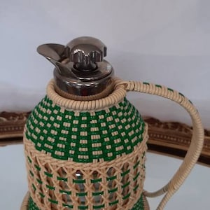 May include: A decorative jug with a woven green and beige pattern. The jug has a silver lid and handle. The design is reminiscent of a vintage style, suitable for serving beverages.