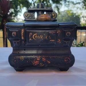 May include: Black ceramic cookie jar with gold accents and the word "Cookies" on the front. The jar has a decorative design on the front and base, and a lid with a handle.
