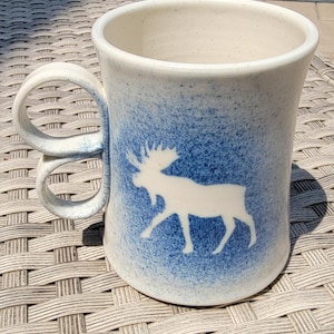 May include: A ceramic mug with a white moose silhouette on a blue speckled background. The mug has a unique double-loop handle and a cream-colored interior. The mug is designed for hot beverages.